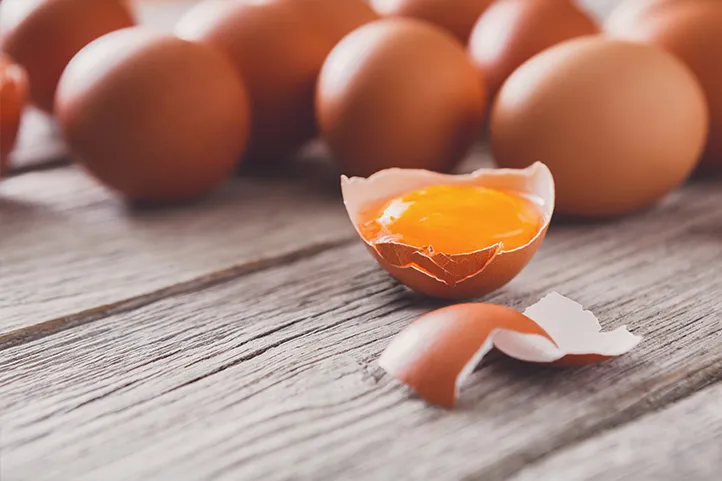 A group of brown eggs placed on a wooden table, with one half shell in the foreground containing a yellow egg yolk