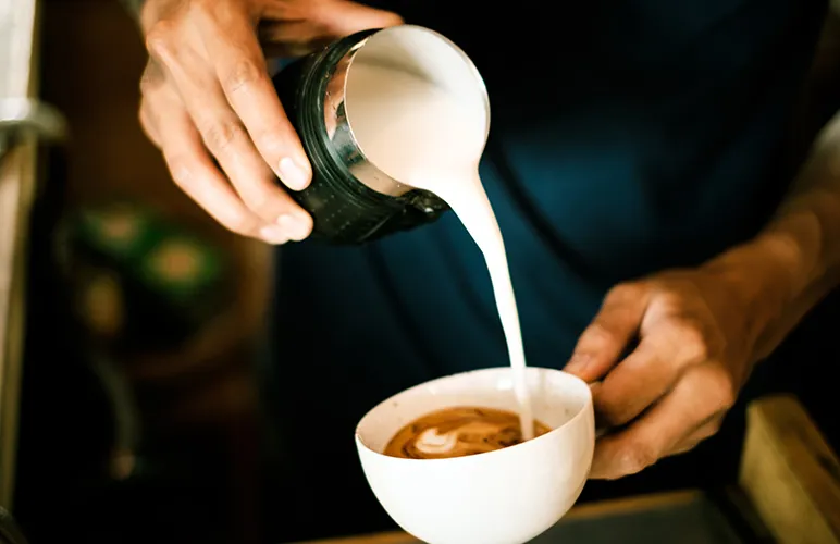 Milk being poured from a metal container into a white cup containing coffee